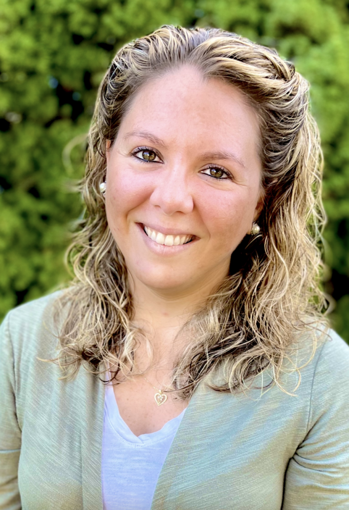 Valerie Tornini smiling in a close-up outdoor portrait with leafy green trees in the background.